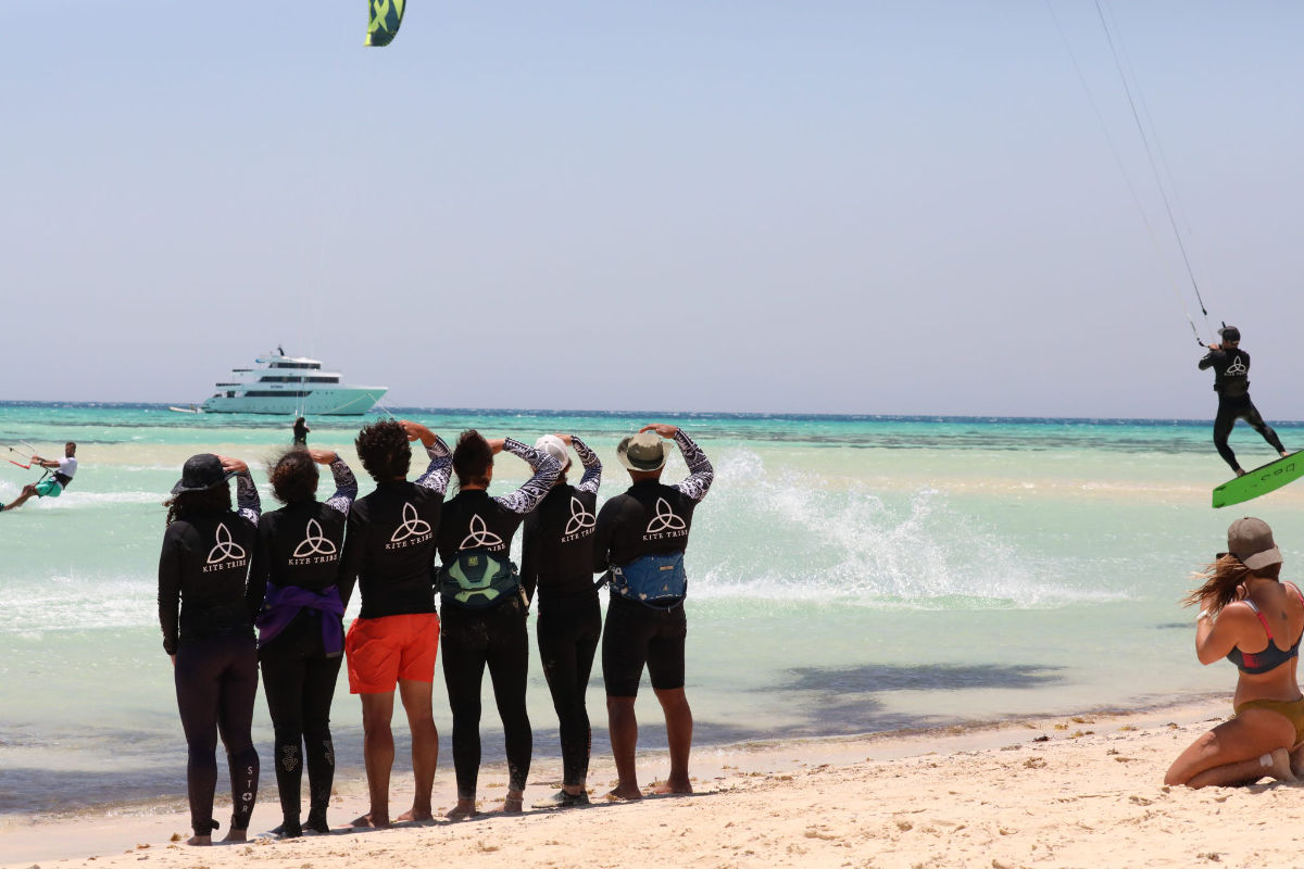 group of people on the beach watching a kitesurfer jump. everyone is wearing a kite tribe tshirt