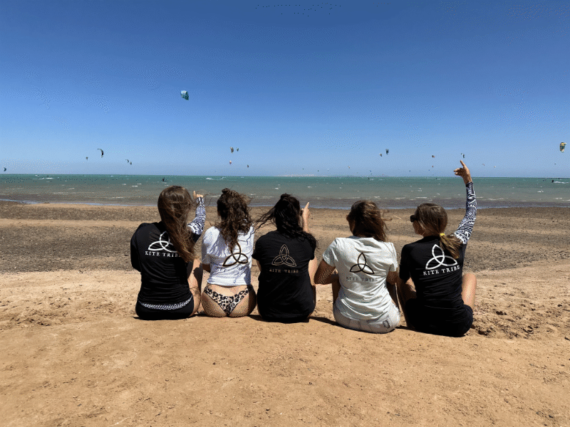 group of 5 girls sitting on the kite beach in El Gouna in Egypt, looking at the kites in the water. The image shows the girls from the back, all wearing a Kite Tribe T-Shirt