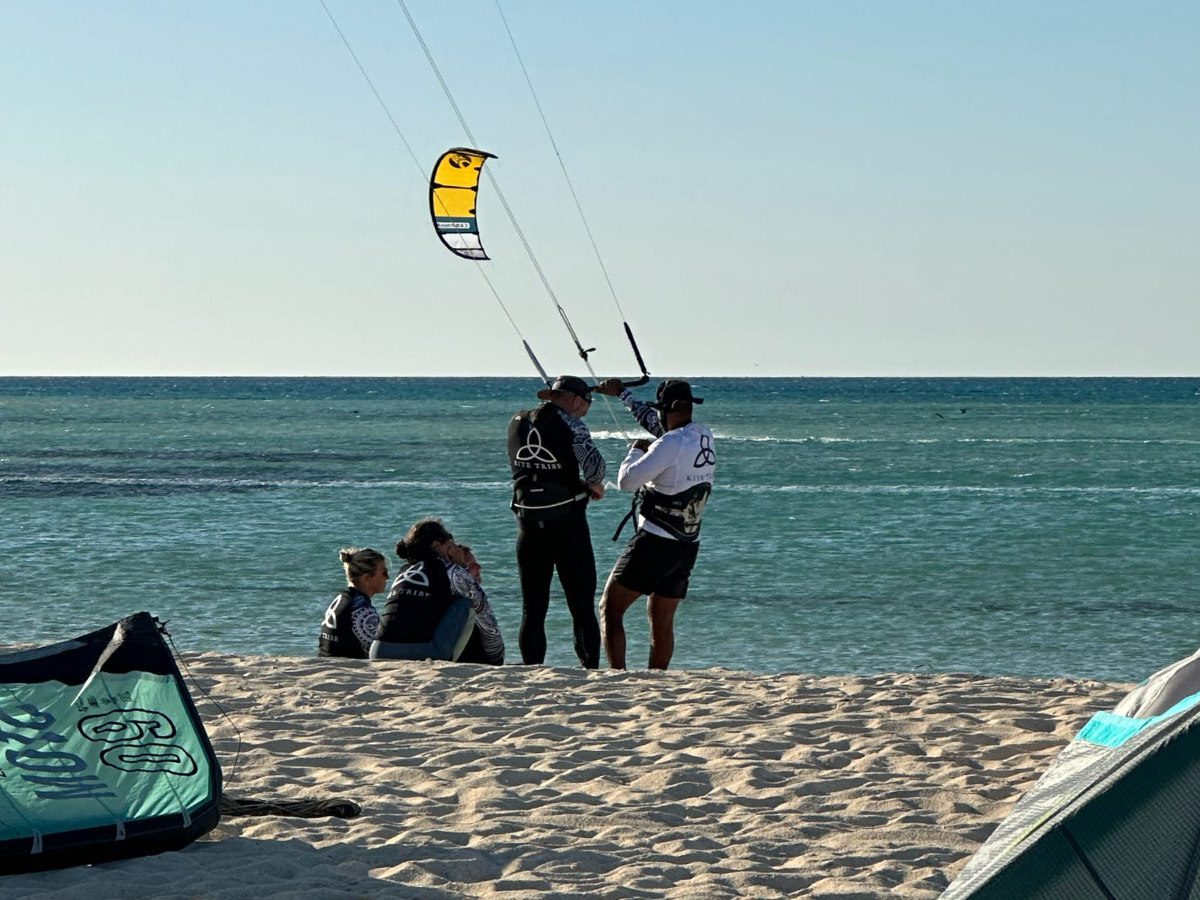 group of 5 girls sitting on the kite beach in El Gouna in Egypt, looking at the kites in the water. The image shows the girls from the back, all wearing a Kite Tribe T-Shirt
