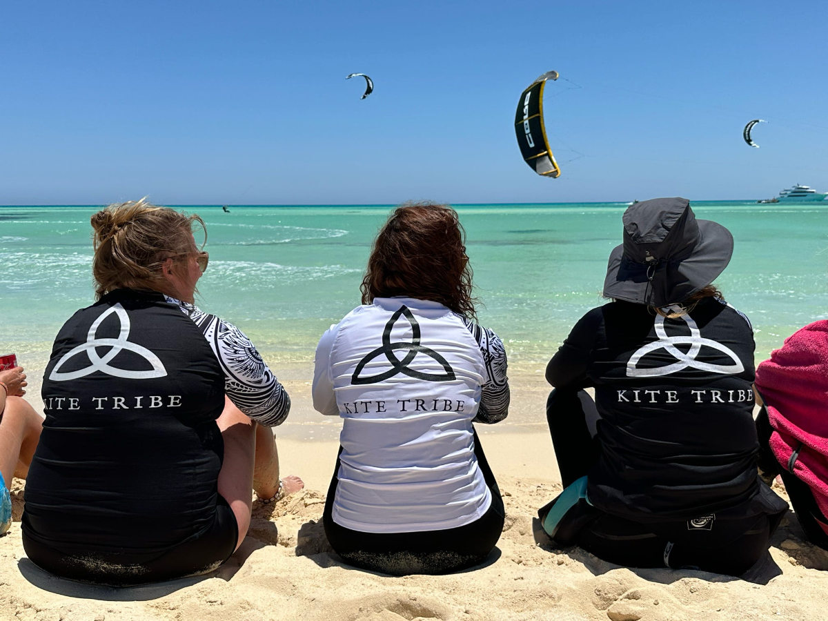 group of 5 girls sitting on the kite beach in El Gouna in Egypt, looking at the kites in the water. The image shows the girls from the back, all wearing a Kite Tribe T-Shirt