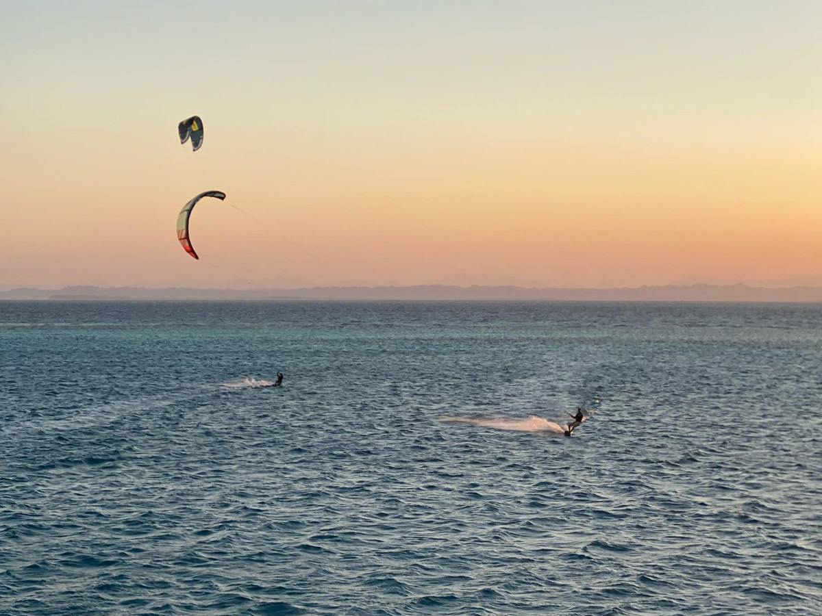group of 5 girls sitting on the kite beach in El Gouna in Egypt, looking at the kites in the water. The image shows the girls from the back, all wearing a Kite Tribe T-Shirt