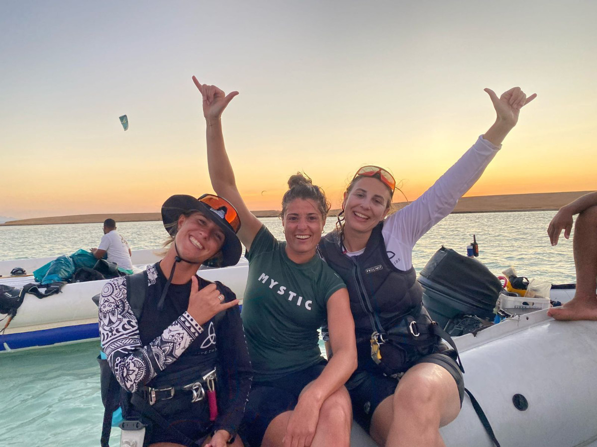 kite-safari--girls-in-zodiac group of 5 girls sitting on the kite beach in El Gouna in Egypt, looking at the kites in the water. The image shows the girls from the back, all wearing a Kite Tribe T-Shirt